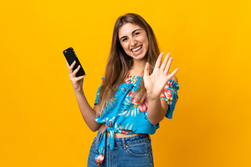 Wall Mural - Young woman using mobile phone over isolated yellow background saluting with hand with happy expression