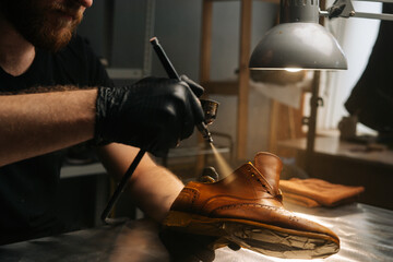 Close-up hands of unrecognizable shoemaker wearing black gloves spraying paint of light brown leather shoes. Concept of cobbler artisan repairing and restoration work in shoe repair shop.