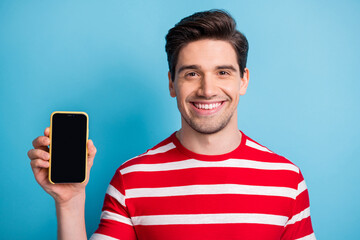 Wall Mural - Photo of positive brunette young man hold phone show screen empty space isolated on blue color background