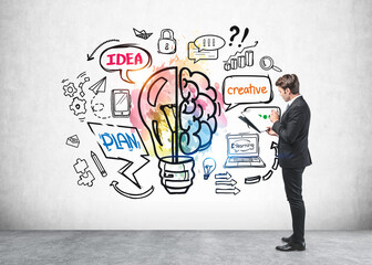 Wall Mural - Side view of businessman in suit checking his notes in folder and standing in front of concrete wall with colorful lightbulb and brain sketch. Concept of success and future career opportunities.