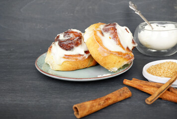 Two cinnabons buns lie on a green plate and next to cinnamon sticks and cane sugar in the right corner on a gray wooden background  