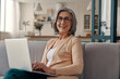 © gstockstudio - Beautiful senior woman in casual clothing using laptop and looking at camera while sitting on the sofa at home