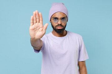 Wall Mural - Young strict serious unshaven student black african man 20s wear violet t-shirt hat glasses do stop palm gesture refusing say no look camera isolated on pastel blue color background studio portrait.