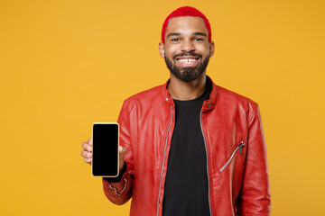 Poster - Young african fun smiling man 20s with funky pink hairdo in red leather jacket hold mobile cell phone blank screen workspace area looking camera isolated on yellow orange background studio portrait.