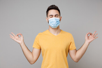 Wall Mural - Young man in yellow t-shirt in sterile face mask to safe coronavirus covid-19, pandemic quarantine hold hands in yoga gesture meditating, try to calm down isolated on grey background studio portrait.