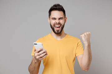 Wall Mural - Young caucasian smiling happy bearded attractive man in casual yellow t-shirt hold mobile cell phone chat in social network do winner gesture clench fist isolated on grey background studio portrait.