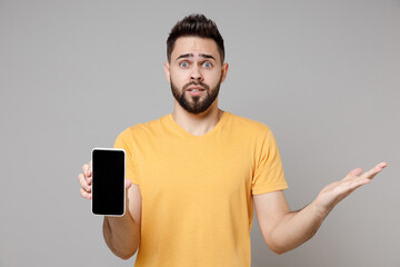 Wall Mural - Young confused shocked bearded caucasian man 20s wear yellow basic t-shirt hold mobile cell phone, blank screen workspace area spreading hand oops gesture isolated on grey background studio portrait.
