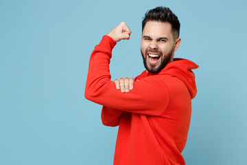 Wall Mural - Young caucasian bearded strong sporty fitness attractive man 20s in casual red orange hoodie showing biceps muscles on hand isolated on blue color background studio portrait People lifestyle concept.