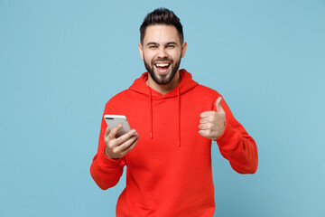 Wall Mural - Young caucasian smiling happy bearded man 20s in casual red orange hoodie hold mobile cell phone texting show thumb up gesture isolated on blue background studio portrait People lifestyle concept