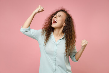 Young black african american happy expressive excited happy positive curly woman 20s in blue shirt do winner gesture clench fist celebrating scream isolated on pastel pink background studio portrait