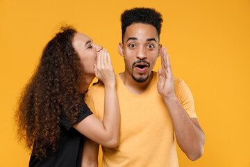 Wall Mural - Young couple two friends family african shocked man woman in black t-shirt whisper gossip and tell secret behind hand share news with guy spreading hand isolated on yellow background studio portrait.