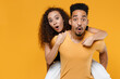 © ViDi Studio - Young couple two friends together family african shocked surprised man woman with open mouth in black t-shirt giving piggyback ride to joyful sit on back isolated on yellow background studio portrait.