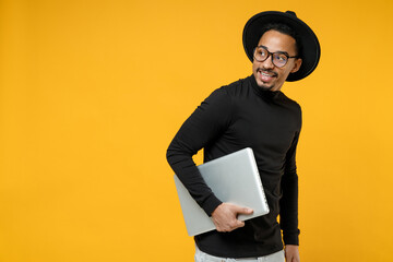 Wall Mural - Side view of young freelancer smiling copywriter african man 20s wear stylish black shirt hat eyeglasses walk with closed laptop pc computer look aside isolated on yellow background studio portrait