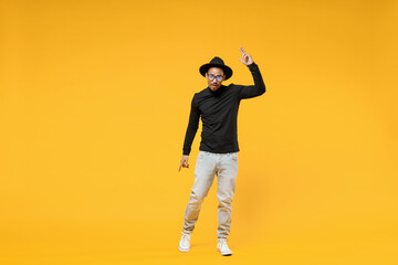 Wall Mural - Full length young fun african man 20s wearing stylish black hat shirt eyeglasses doing winner gesture pointing index finger up look camera isolated on yellow orange color background studio portrait