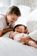 © Sergio Marcos/Stocksy - Cheerful parents lying on bed with newborn baby in cozy sunny bedroom