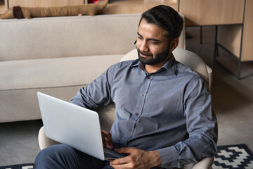 Serious bearded indian business man working on laptop from home office sitting in chair. Male indian entrepreneur using computer remote studying, browsing web, having virtual meeting, watching webinar