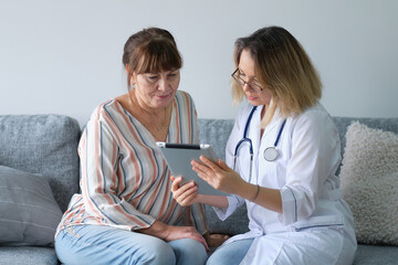 Female professional doctor showing medical test result explaining prescription using digital tablet app visiting senior woman patient at home sitting on sofa. Elderly people healthcare tech concept.