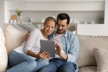 Happy young man and woman spouses relax on sofa in living room using modern tablet gadget together. Smiling Caucasian couple rest on couch at home talk on video call on pad. Technology concept.