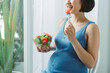 © Maki Company Limited/Stocksy - Happy pregnant woman enjoying a salad sitting on the sofa at home