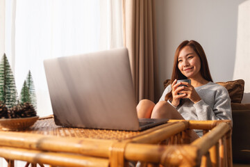 A beautiful young asian woman using and working on laptop computer while drinking coffee at home