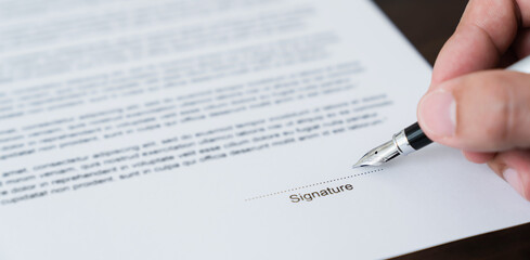 Hand holds pen and signs agreement contract on the table.