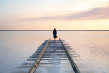 Woman Standing On Railway Going Into A Pink Lake At Sunset