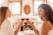 Charme, beauty and happiness: multiracial young women couple toasting with red wine looking in the eyes each other with bright sun entering the house through the open windows illuminating the room