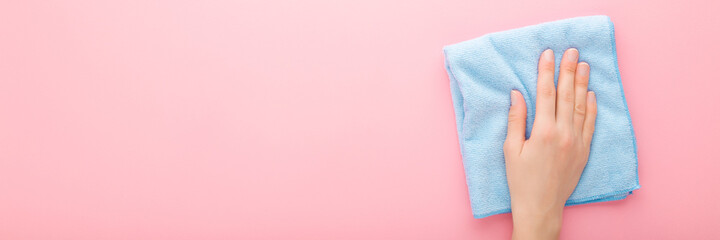 Young adult woman hand holding blue rag and wiping table, wall or floor surface in kitchen, bathroom or other room. Closeup. Pastel pink background. Wide banner. Empty place for text. Top down view.