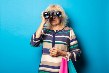 Old woman holding shopping bags and looking through binoculars on blue background