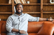 © Vadim Pastuh - Ethnic black unshaved plump man with dreadlocks sitting on the leather sofa and smiling, in a normal blue shirt and jeans, feeling comfort and relaxation, at an interview, bookcase in the background