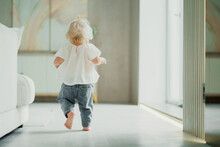The Child Plays Running Around At Home Alone. Children And Home. A Cheerful Mood. Portrait Of A Cute Little Girl With Blonde Blue Eyes. The Family Spends Time At Home On The Couch.