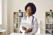 © Studio Romantic - Portrait of happy professional doctor at work. Young black woman in white lab coat with stethoscope standing in medical office or exam room at clinic, holding clipboard, smiling and looking at camera