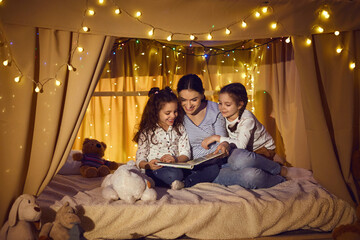 Wall Mural - Happy family reading good book in the evening. Young mother telling bedtime stories to little children. Mommy and daughters enjoying fairy tales sitting in cozy playroom tent decorated with LED lights