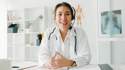 Young Asia lady doctor in white medical uniform with stethoscope using computer laptop talk video conference call with patient, looking at camera in health hospital. Consulting and therapy concept.