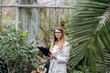 © sofiko14 - Young high-skilled woman agronomist wearing white lab coat, walking with folder beside palm trees in beautiful greenhouse, making notes and supervising plants condition