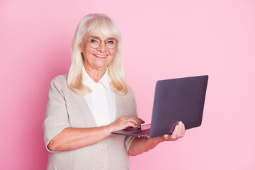 Wall Mural - Photo of cheerful old woman hold netbook wear eyewear beige cardigan isolated pastel pink color background