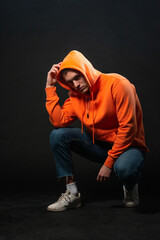 Wall Mural - Male model sitting on the floor and posing in orange sports hoodie isolated over a black background.