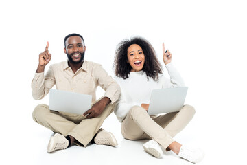 excited african american couple pointing up with fingers while sitting with laptops on white