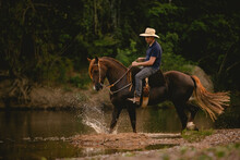 White Man Riding A Horse Inside A River, Wearing A Straw Hat, With His Horse In A Saddle And Throwing Water Into The Air.