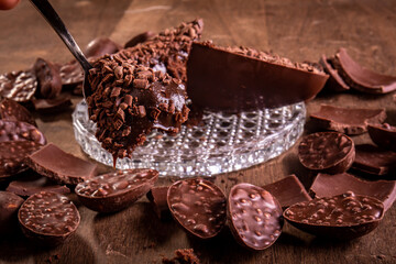 Spoonful of a cracked stuffed chocolate easter egg with grated chocolate on the top and lots of mini easter eggs and chocolates all around it on a glass stand on a wooden table.