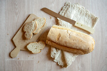 High Angle Close Up Of Freshly Baked White Bread, Bread Knife, Cutting Board And Linen Napkin On Wooden Table.