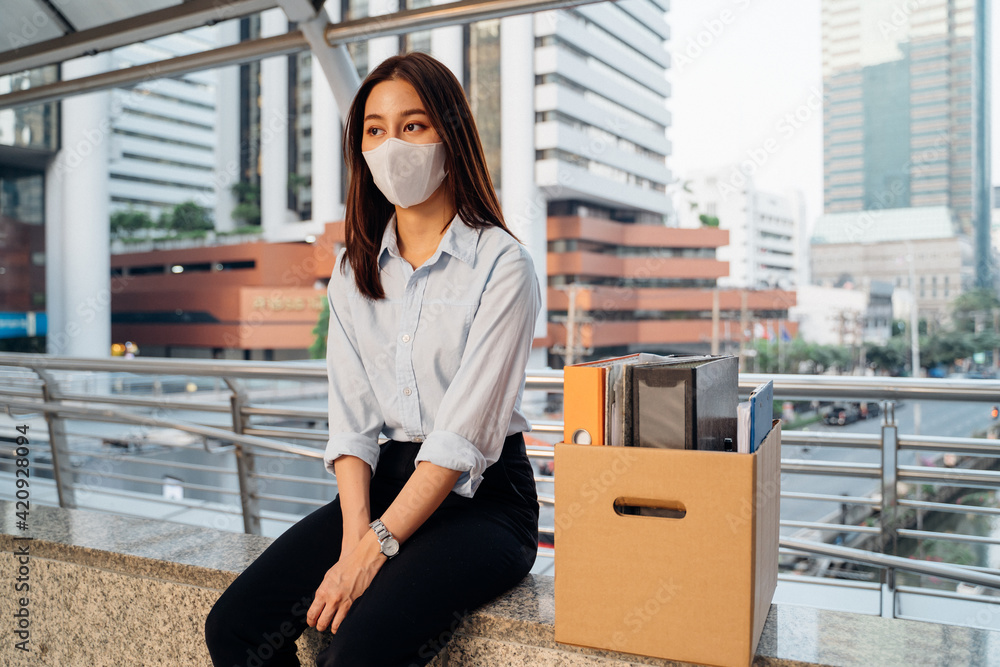 Stressed and worried young Asian woman with box of items sitting alone after being laid off from job due to covid-19 pandemic - obrazy, fototapety, plakaty 