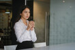 © Prathankarnpap - Young woman office worker holding coffee cup or tea and looking away thoughtfully.