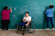 © Bangkok Click Studio - Young Asian disabled boy without legs sitting on a wheelchair, smile and looking to camera while his friend fun with playing and writing on green wall in classroom. Idea for handicapped kid studying