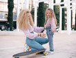 © vera - Mother and daughter sit on skateboard. Woman with unusual, lush and long hair. Small girl and woman smiles and happy. same colour  of clothes, get fun outdoor in weekend. Small kid study skating.