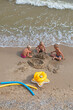 © Nataliia Makarovska - happy tanned children building a sand castle by the sea with rubber duck and banana on the beach on a summer day, vertical format