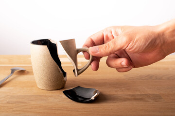 on a slatted wooden table is a broken brown coffee cup. The hand of a man brings the piece of the handle closer together making a kind of puzzle