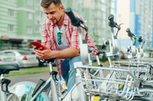 Positive Man Stands By The Parking Lot With A Bicycle Rental And Uses A Smartphone With A Smile On His Face. Tourist Shares A Bicycle For A Walk On The Internet With A Smartphone