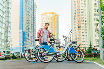 Handsome man in a shirt stands with a bicycle in the bicycle rental parking lot and looks at the vehicle with a serious face. 