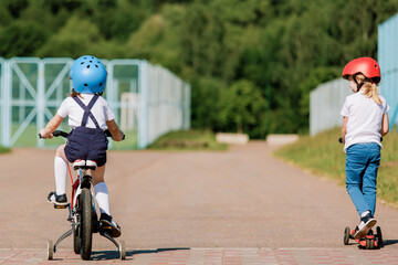 Backview of two little girls having fun on bicycle and scooter. Cheerful sisters in helmets riding outdoors.